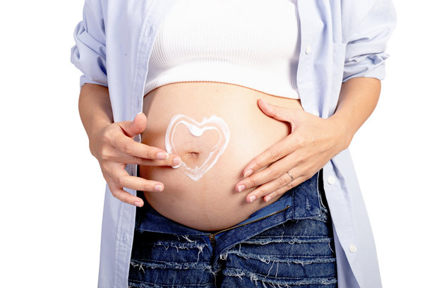 快乐的亚洲孕妇在白色背景下，母亲正在给她的大肚子涂抹保湿霜。妈妈用化妆品在肚子上画心形图案。图片下载