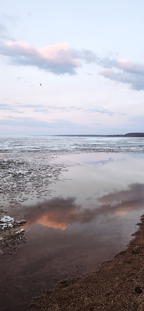 春季风景。美丽的海湾晨曦全景。明亮的光线下，水面上漂浮着积云。海岸线上有冰雪和岩石。阳光的光芒在海面上反射。竖版照片图片下载