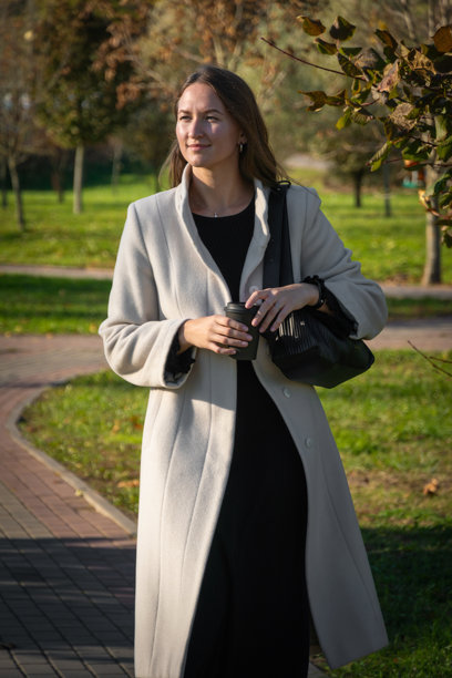 年轻女子穿着轻便外套，手握咖啡杯，在公园里漫步，享受美丽秋日的休闲时光。竖版照片图片下载