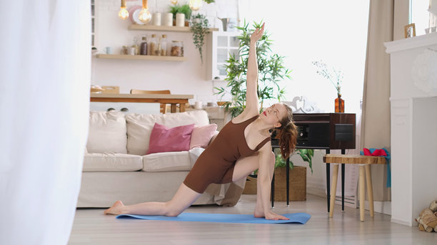 一位穿着棕色背心和短裤的女性在明亮的客厅里，平衡在蓝色瑜伽垫上进行拉伸。这张照片捕捉了家庭健身和追求身体健康的本质。图片下载