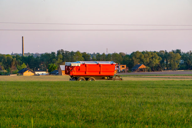 红色农业拖车停在绿色田野中，背景是建筑和森林。地平线上可以看到烟囱和一排树木。照片是在日落时拍摄的，呈现出温暖的色调。图片下载