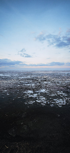 春季风景。美丽的黎明全景，海湾上方的积云在明亮的光线下。海岸线上有冰、雪和岩石。阳光的光芒在海面上反射。竖版照片图片下载