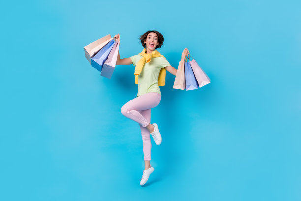 乐观的年轻棕发女士穿着T恤、裤子和运动鞋，手提购物袋在蓝色背景下跳跃的全身照片图片下载
