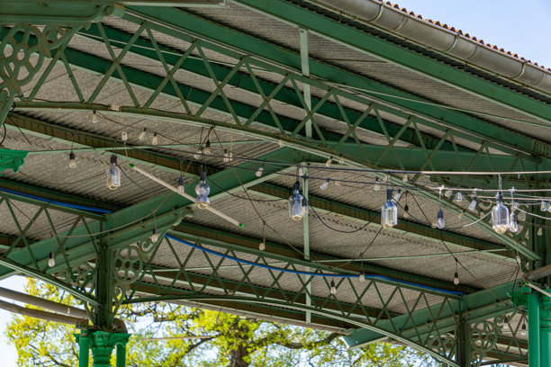 工业风格屋顶的钢结构特写，涂成绿色，屋顶悬挂着装饰灯和灯泡。背景中有树木和建筑的屋顶。照片拍摄于阳光明媚的日子。图片下载