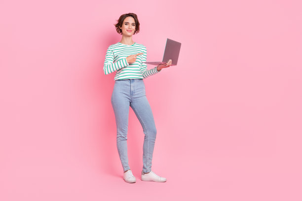 穿着衬衫、牛仔裤和鞋子的甜美千禧一代短发女士，坐在粉色背景前，手指着笔记本电脑的全身照片。图片下载