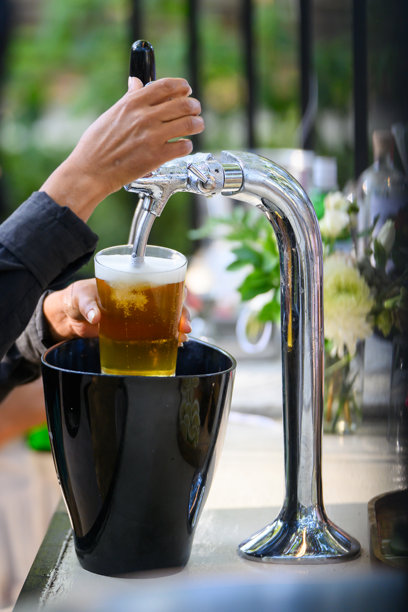 金色啤酒从光滑的金属水龙头倒入玻璃杯中，置身户外。图片下载