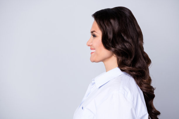 乐观的棕发波浪年轻女性穿着蓝色服装，背景为灰色，个人资料照片。图片下载