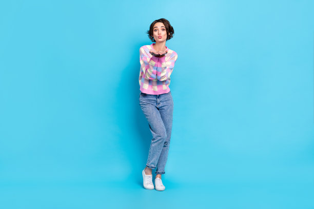 可爱的年轻棕发女孩穿着毛衣、牛仔裤和运动鞋，吹吻的全身照片，背景为蓝色。图片下载