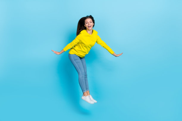可爱的千禧世代棕发女士穿着衬衫和牛仔裤，跳跃在蓝色背景上，照片为全尺寸。图片下载