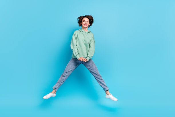 可爱的年轻棕发女孩穿着毛衣、牛仔裤和鞋子在蓝色背景上跳跃的全身照片图片下载