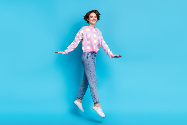 可爱的千禧世代棕发女孩穿着毛衣、牛仔裤和鞋子在蓝色背景上跳跃的全身照片图片下载