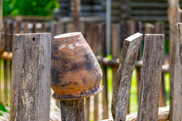 传统陶土壶放置在乡村环境中的木栅栏上。照片展示了乡村风景和手工艺的元素，适合用于阐述与传统和民俗文化相关的话题。图片下载