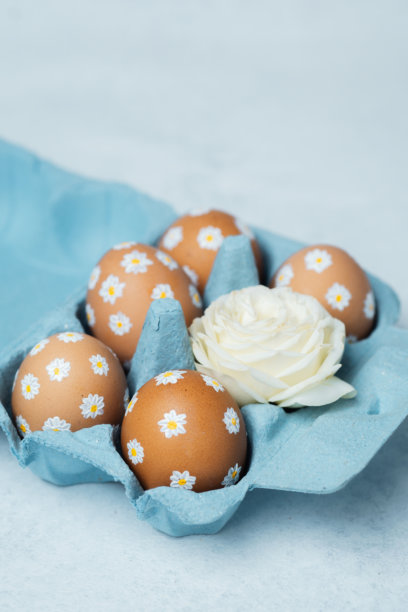 手绘的复活节彩蛋，带有雏菊图案，放在蓝色纸箱中，旁边有白色玫瑰。复活节主题的构图，背景为浅色。图片下载