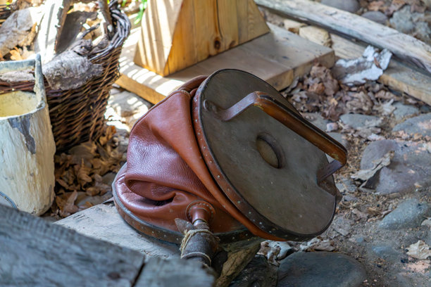 古老的木质和皮革制成的铁匠风箱，躺在木材和石头之间。照片展示了一种传统的手工工具，置于乡村自然环境中。非常适合用于阐述与古老工艺相关的主题。图片下载