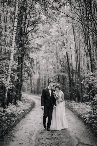 一张黑白照片，展示了一对新婚夫妇手牵手走在森林小径上，微笑着亲吻， bride手中捧着浪漫的花束。图片下载