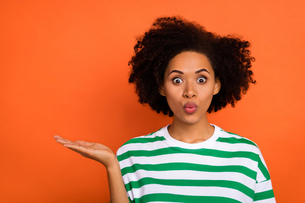 一位迷人的时尚波浪卷发女孩手掌托着空白空间，展示惊艳优惠，背景为明亮的橙色。图片下载