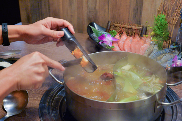 一个人正在往冒着热气的火锅中添加食物，旁边有海鲜和蔬菜。图片下载