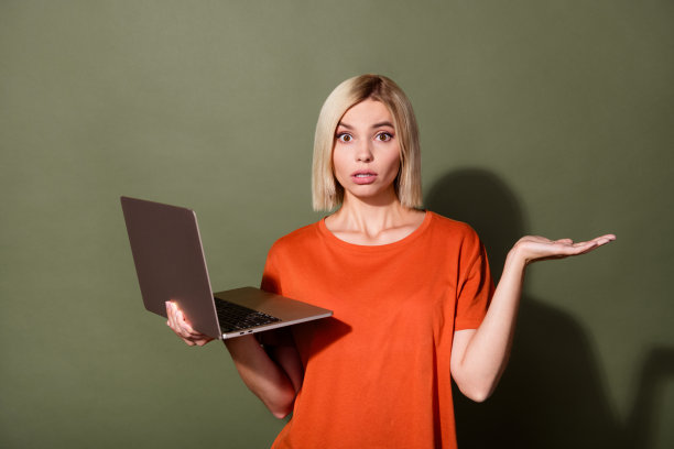 漂亮年轻女孩的照片肖像，手持笔记本电脑，神情困惑和担忧，穿着时尚的橙色服装，背景为卡其色。图片下载