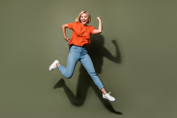 年轻金发女性在卡其色背景上跳跃的全身照片，展现出强壮的二头肌，运动教练风格。图片下载