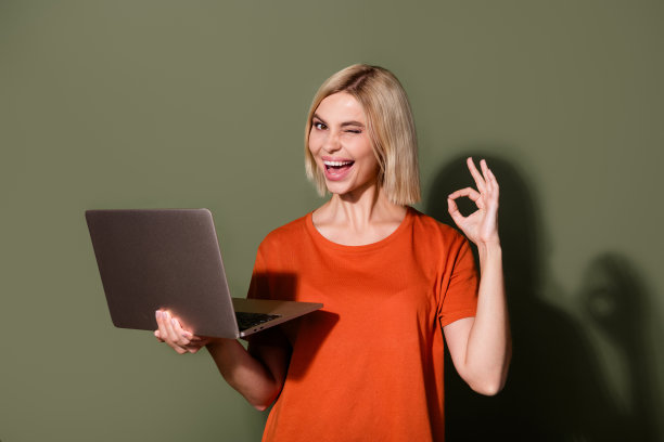 漂亮年轻女孩的照片肖像，手持笔记本电脑，做出“OK”手势，眨眼，穿着时尚的橙色服装，背景为卡其色。图片下载