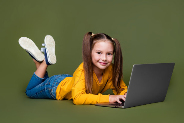 聪明可爱的小孩穿着黄色毛衣和牛仔裤，躺在卡其色背景上用笔记本电脑做作业的全身照片。图片下载