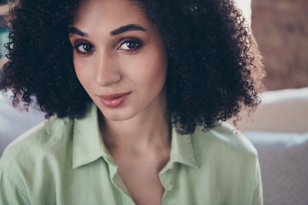 裁剪的照片，展示了一位可爱的女士，卷曲的秀发，甜美的面容，穿着卡其色衬衫，背景是和谐宁静的室内客厅。图片下载