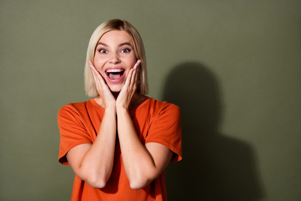 漂亮年轻女孩的照片肖像，双手托着脸颊，兴奋地欣赏自己，穿着时尚的橙色服装，背景为卡其色。图片下载