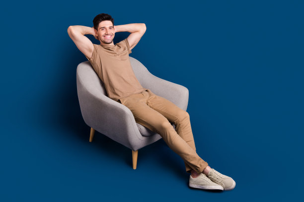 穿着米色T恤的积极心态男士在蓝色背景下的扶手椅中放松，享受周末时光的全身照片图片下载