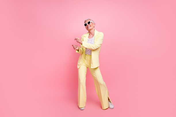 穿着节日黄色西装的无忧无虑的舞动的老年女商人全身照片，背景为粉色。图片下载