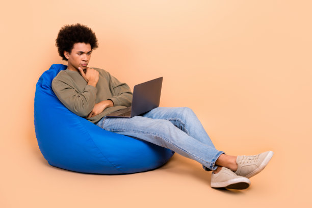 年轻的男性开发者，身穿智能团队领导IT专家的全身照片，坐在豆袋椅上，手托下巴，使用笔记本电脑，背景为米色。图片下载