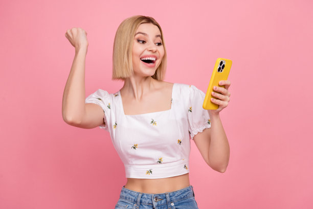 疯狂女孩的照片，金发，穿着上衣，举起拳头，手持智能手机，欢呼庆祝，赢得宇宙乐透彩票，背景为粉色。图片下载