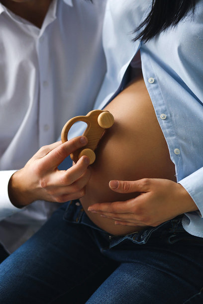 怀孕的女人穿着蓝色衬衫和牛仔裤，丈夫在她的孕肚上放着一个木制儿童玩具车的特写照片，室内家庭概念。图片下载