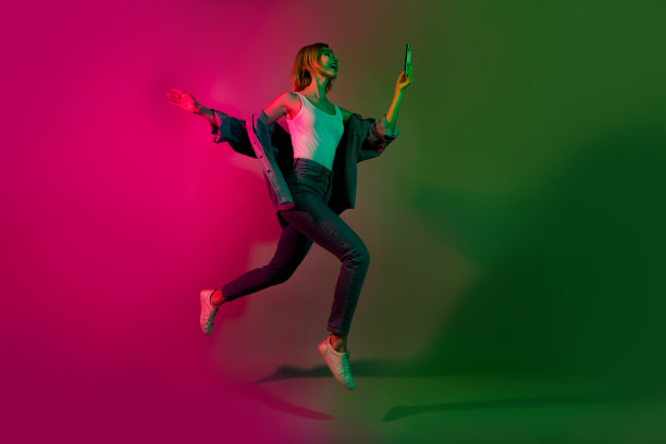 全身照片，快乐的女士在迪斯科活动中快速跳跃走动，手持智能设备发送短信，背景为鲜艳的霓虹色。图片下载