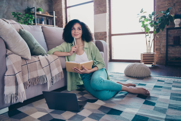 全身照片，迷人的赤脚女孩坐在地毯上，手持笔记本和钢笔，使用笔记本电脑，沉思于公寓的阁楼室内。图片下载