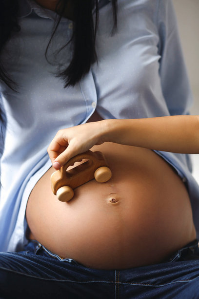 孕妇穿着蓝色衬衫和牛仔裤，女儿手握木制儿童汽车玩具，靠在母亲的孕肚上，室内特写照片。家庭概念。图片下载