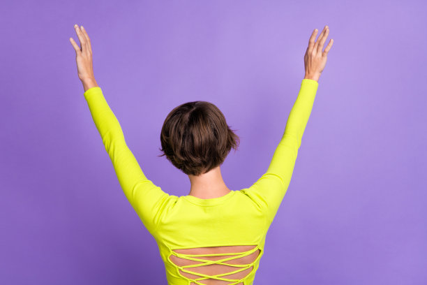背面视角照片，搞笑的女性人物，棕色短发，穿着时尚的短款上衣，双臂高举，背景为紫色。图片下载