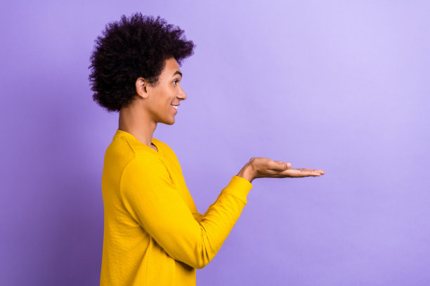 侧面照片，帅气的男士穿着时尚的黄色毛衣，手臂伸出，似乎在提供什么，背景为紫色，显得空旷。图片下载