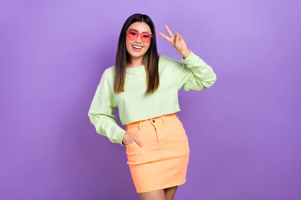 穿着绿色卫衣、戴着粉色眼镜的可爱兴奋女孩在紫色背景前做出V字手势的照片图片下载