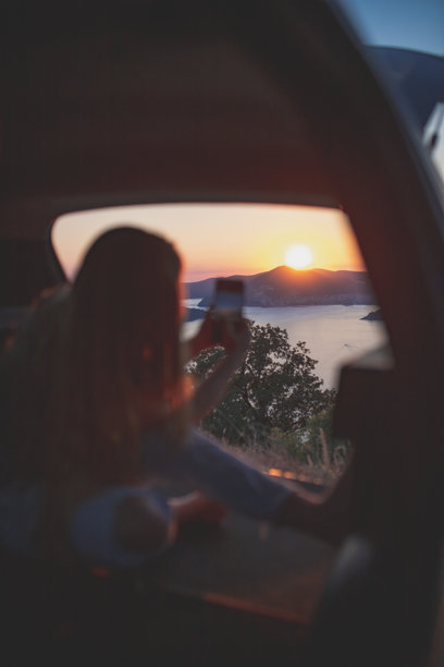 年轻女性坐在汽车后备箱里拍摄日落照片图片下载