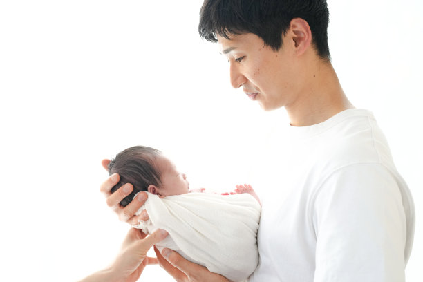 一位二十多岁的父亲抱着他七天大的日本新生儿，站在白色背景前。这张图片捕捉了父亲与孩子之间温馨的爱与联系时刻。图片下载