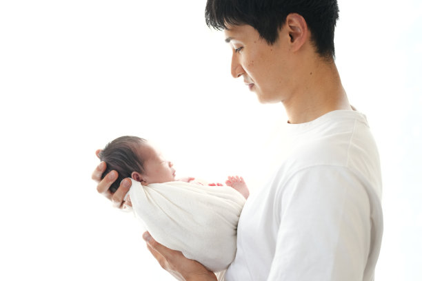 一位二十多岁的父亲抱着他七天大的日本新生儿，站在白色背景前。这张图片捕捉了父亲与孩子之间温馨的爱与联系时刻。图片下载