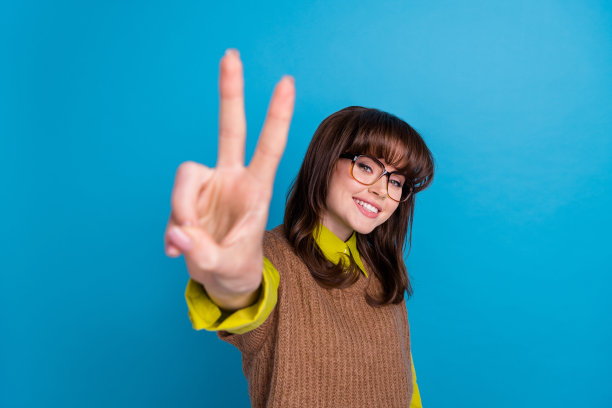 穿着棕色背心、戴着眼镜的活泼快乐女士在蓝色背景前做出V字手势的照片图片下载