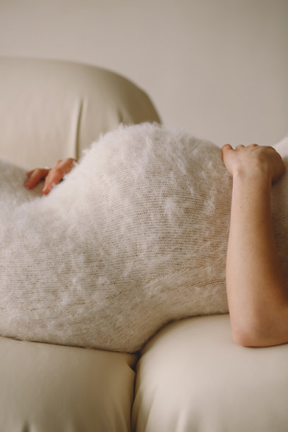 穿着浅米色毛茸茸裙子的孕妇躺在沙发上，手捂着肚子。孕肚特写照片。图片下载
