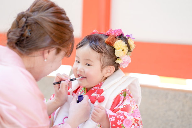 一个穿着红色和服的三岁日本女孩在庆祝七五三。她的妈妈穿着粉色和服，为她化妆。这张照片展示了母女在这个特殊日子里的温馨时刻。图片下载