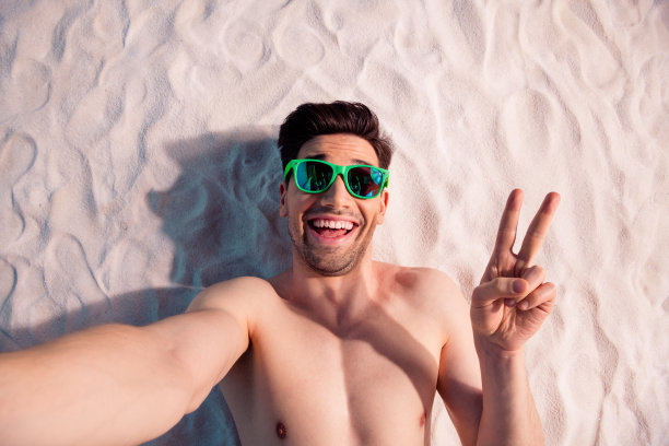 俯视照片中，积极向上的时尚男士穿着泳装，躺在沙滩上，做出V字手势，正在户外海滩录制视频博客。图片下载