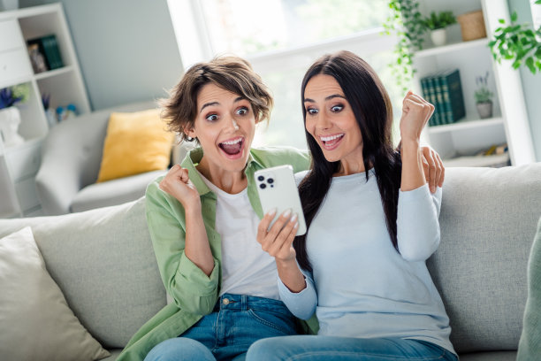 年轻女孩们穿着衬衫，坐在沙发上，兴奋地庆祝在现代设备上赢得游戏，室内公寓房间的照片。图片下载
