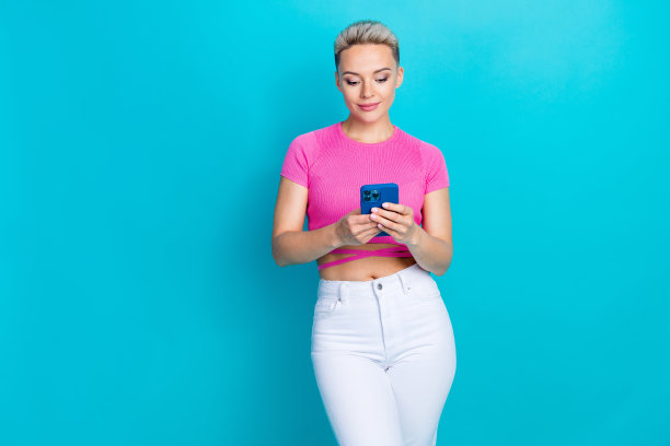 漂亮年轻女孩的照片肖像，手持电话阅读新闻并发表评论，穿着时尚的粉色服装，背景为青色。图片下载