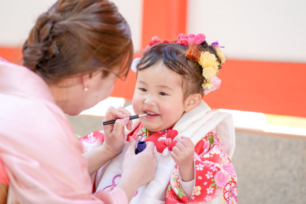 一个穿着红色和服的三岁日本女孩在庆祝七五三。她的妈妈穿着粉色和服，为她化妆。这张照片展示了母女在这个特殊日子里的温馨时刻。图片下载