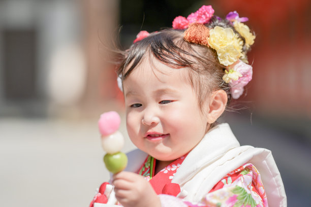 一个穿着红色和服的三岁日本女孩在庆祝七五三。她坐着吃着妈妈给的团子，妈妈穿着粉色和服。这张照片捕捉了母女之间甜美的瞬间。图片下载