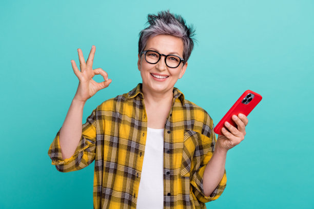 迷人的祖母照片肖像，手持电子设备，做出“OK”手势，穿着时尚的黄色格子服装，背景为青色。图片下载